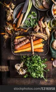 Organic root vegetables in harvest basket on dark rustic kitchen table background with ingredients for tasty cooking with greens flavor and kitchen tools, top view. Healthy clean food and eating
