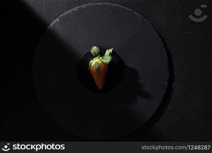 Organic strawberry on black platter in harsh shadow on black background. Above view of one ripe strawberry. Sweet summer fruit. Single red berry.
