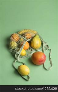 Organic tropical fruits mix in a fabric mesh shopping bag on a green background. Food shopping concept. Summer exotic fruits. Fruit retail context.