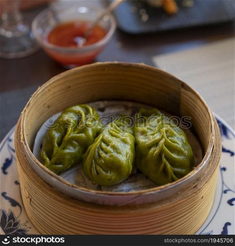 Oriental green dumplings in a traditional bamboo steamer, background chili sauce at restaurant.