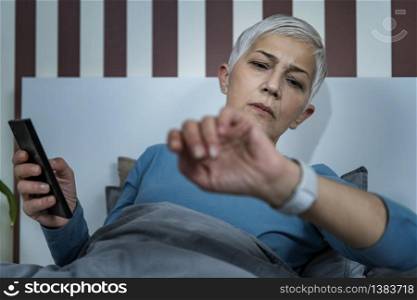 Orthosomnia Sleeping Disorder ? Woman in Bedroom, Using Smart Phone and Watch for Sleep Tracking, Concerned about Her Sleeping Habits