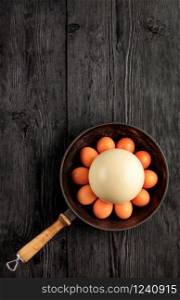 Ostrich egg, selective focuse, surrounded by chicken eggs in an old cast-iron skillet, which is standing on an old black wooden surface, top view, copy space, vertical image.. Ostrich egg, selective focuse, surrounded by chicken eggs in an old cast-iron skillet, vertical image.