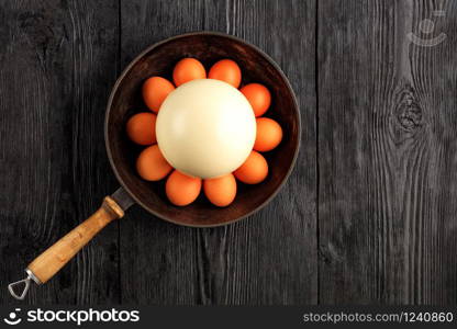 Ostrich egg surrounded by chicken eggs in an old cast-iron skillet, which is standing on an old black wooden surface, top view, copy space.. Ostrich egg surrounded by chicken eggs in an old cast-iron skillet.