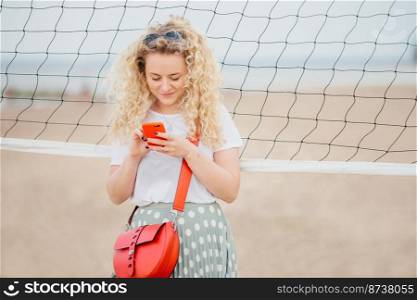 Oudoor shot of pleasant looking blonde female holds cell phone, shares photos in social networks, dressed casually, uses free internet, has walk across beach, stands near tennis net. Technology