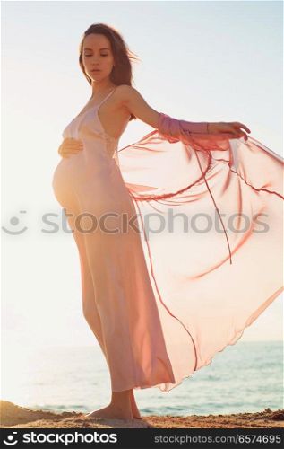 Outdoor atmospheric lifestyle portrait of beautiful pregnant woman in pink dress. Beautiful morning. Sunrise on the beach. Beauty and fashion