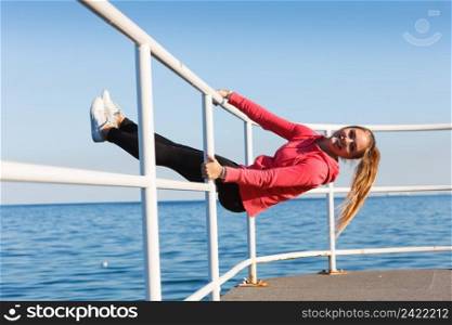 Outdoor, dangerous play, sport concept. Woman in sports suit sitting on handrail next to sea and having fun while exercising.. Woman fool around sitting on handrail near sea