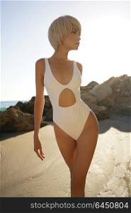 Outdoor fashion photo of graceful young woman in white swimwear on the beach. Getting golden tan. Fashion and beauty. Summer travel