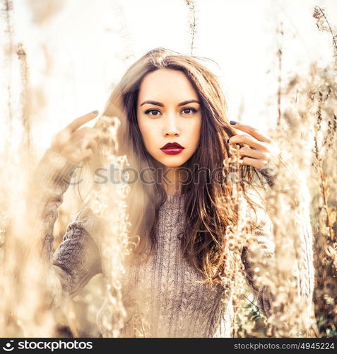 Outdoor fashion photo of young beautiful lady in autumn landscape with dry flowers. Knitted sweater, wine lipstick. Warm Autumn. Warm Spring