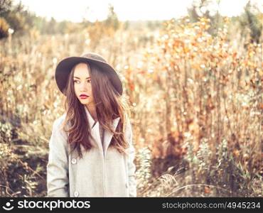 Outdoor fashion photo of young beautiful lady in autumn landscape with dry flowers. Gray coat, black hat, wine lipstick. Warm Autumn. Warm Spring