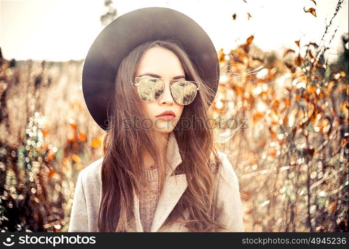 Outdoor fashion photo of young beautiful lady in autumn landscape with dry flowers. Gray coat, black hat, sunglusses, wine lipstick. Warm Autumn. Warm Spring