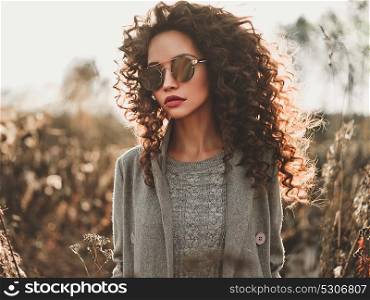 Outdoor fashion photo of young beautiful lady in autumn landscape with dry flowers. Gray coat, knitted sweater, sunglasses, wine lipstick. Fashion lookbook. Warm Autumn. Warm Spring