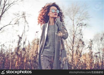 Outdoor fashion photo of young beautiful lady in autumn landscape with dry flowers. Gray coat, knitted sweater, sunglasses, wine lipstick. Fashion lookbook. Warm Autumn. Warm Spring
