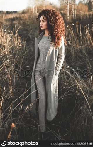 Outdoor fashion photo of young beautiful lady in autumn landscape with dry flowers. Gray coat, knitted sweater, wine lipstick. Fashion lookbook. Warm Autumn. Warm Spring