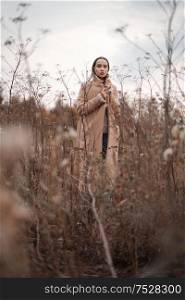 Outdoor fashion photo of young beautiful lady in autumn landscape with dry flowers. Knitted sweater, beige coat. Warm Autumn. Warm Spring