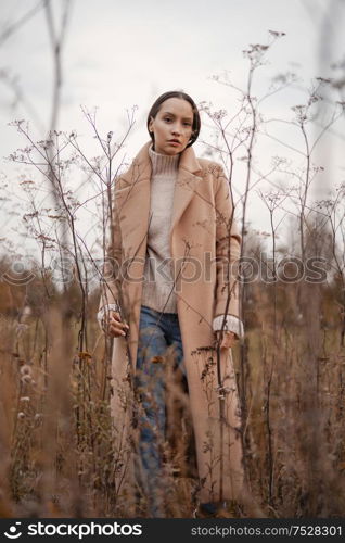 Outdoor fashion photo of young beautiful lady in autumn landscape with dry flowers. Knitted sweater, beige coat. Warm Autumn. Warm Spring