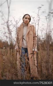 Outdoor fashion photo of young beautiful lady in autumn landscape with dry flowers. Knitted sweater, beige coat. Warm Autumn. Warm Spring