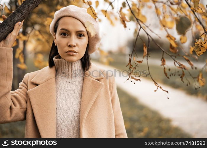 Outdoor fashion photo of young beautiful lady in beige coat, knite sweater and beret surrounded autumn leaves. Warm autumn