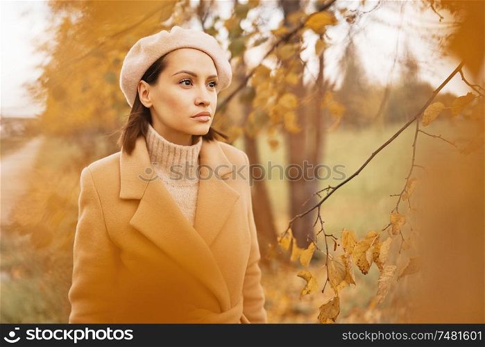 Outdoor fashion photo of young beautiful lady in beige coat, knite sweater and beret surrounded autumn leaves. Warm autumn