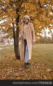 Outdoor fashion photo of young beautiful lady in beige coat, knite sweater and blue jeans in autumn landscape. Warm autumn