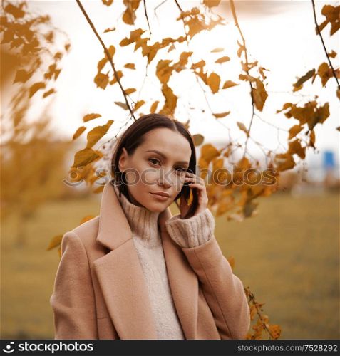 Outdoor fashion photo of young beautiful lady in beige coat surrounded autumn leaves