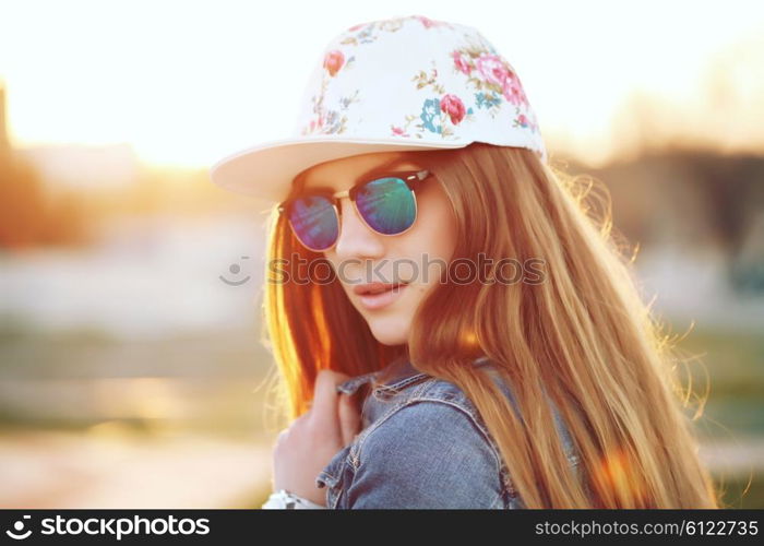 Outdoor fashion portrait of stylish swag girl, wearing swag cap, trendy sunglasses and denim jacket, amazing view of the beach at sunset. Lifestyle picture toned with a vintage instagram filter effect