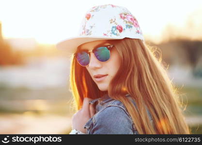 Outdoor fashion portrait of stylish swag girl, wearing swag cap, trendy sunglasses and denim jacket, amazing view of the beach at sunset. Lifestyle picture toned with a vintage instagram filter effect