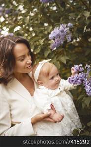 Outdoor fashion portrait of young beautiful mother and little cute daughter in blossoming park. Lilac blossom. Spring image