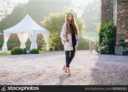 Outdoor horizontal portrait of pleasant looking fashionable woman on high heeled shoes, looks pensively down, poses at camera, stand against wonderful landmarks and white tent. Beauty and nature