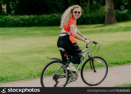 Outdoor image of happy woman rides bike on path, green park around, wears sunglasses, t shirt, pants and sportshoes, has summer holiday, spends spare time actively. People, healthy lifestyle