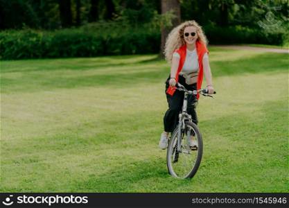 Outdoor image of pretty cheerful young woman rides bike, wears sunglasses, casual wear, poses on green lawn, spends free time in park, bikes in beautiful nature. Activity and recreation concept