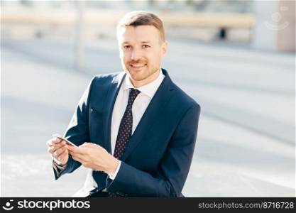 Outdoor image of successful male entrepereneur, wears formal black suit, white shirt with tie, holds smart phone and checks notification on street, sends text message or checks email box online