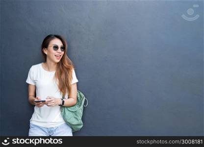 Outdoor lifestyle portrait of pretty sexy young asian girl in travel and glasses style on gray wall background. Asian woman texting message on phone. Copyspace available. Lifestyle and technology.