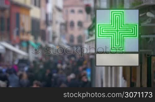 Outdoor pharmacy banner with led green cross hanging on the building. Defocused crowd of people walking in the street in background