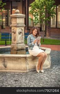 Outdoor photo of beautiful woman reading book on tablet on side of fountain