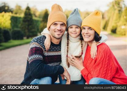 Outdoor portait of beautiful brunette female, handsome man, pretty small girl embrace together, have pleasant mood, walk in park, enjoy calm atmosphere and splendid sunny weather. Friendly family