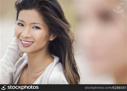 Outdoor portrait of a beautiful happy smiling Chinese Asian young woman or girl at beach with blond female friend out of focus in foreground