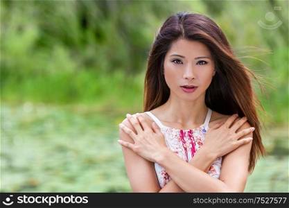 Outdoor portrait of a beautiful young Chinese Asian young woman or girl with perfect make up arms crossed over her chest in front of a natural green background