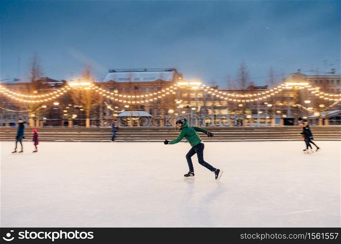 Outdoor portrait of active sporty bearded male involved in winter sport activities, prepares for skating competitions on Christmas ice ring decorated with garlands. Active lifestyle concept.