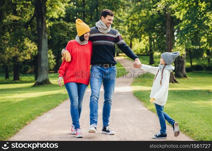 Outdoor portrait of affectionate family walk in park, wear warm knitted clothes. Handsome young man holds daughter`s hand, looks with happy expression at her. Rest and relaxation concept