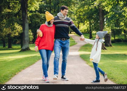 Outdoor portrait of affectionate family walk in park, wear warm knitted clothes. Handsome young man holds daughter`s hand, looks with happy expression at her. Rest and relaxation concept