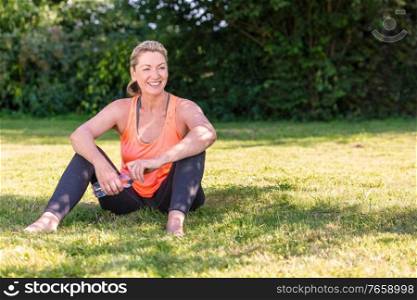 Outdoor portrait of an attractive middle aged blonde woman smiling drinking water relaxing after exercising, running, yoga or fitness lifestyle