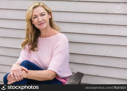 Outdoor portrait of an attractive middle aged blonde woman smiling sitting on a wooden garden chair