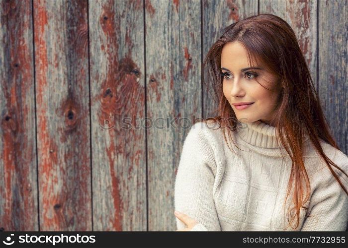 Outdoor portrait of beautiful girl or young woman with red hair wearing a white jumper leaning against an old wooden background