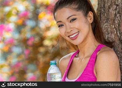 Outdoor portrait of beautiful happy fit healthy Chinese Asian young woman or girl laughing with perfect teeth relaxing after sport with bottle of water in front of natural flower background