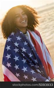 Outdoor portrait of beautiful happy mixed race African American girl teenager female young woman wrapped in the stars and stripes US flag on a beach smiling laughing with perfect teeth in golden sunset evening sunshine