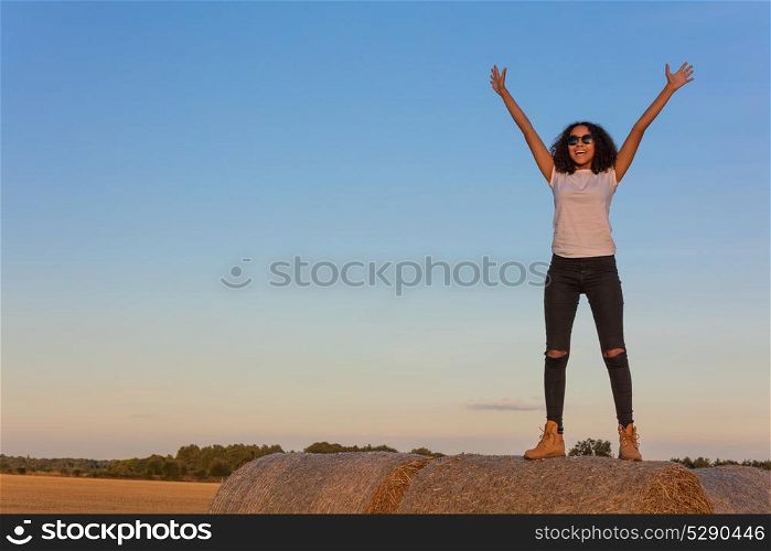 Outdoor portrait of beautiful happy mixed race African American girl teenager female young woman standing celebrating arms raised smiling laughing with perfect teeth and sunglasses on hay bale in golden evening sunshine