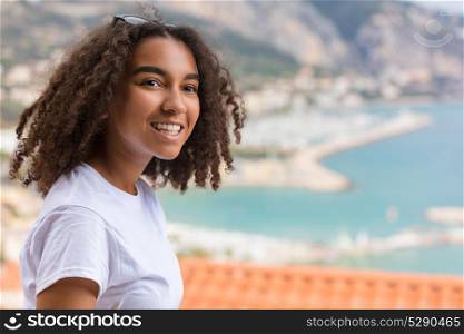 Outdoor portrait of beautiful happy mixed race African American girl teenager female young woman smiling with perfect teeth on vacation with harbor and coastline behind her