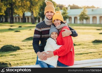 Outdoor portrait of beautiful smiling woman, handsome man and their little cute daughter stand together against ancient bilduing in park, wear warn knitted clothes, have interesting excursion