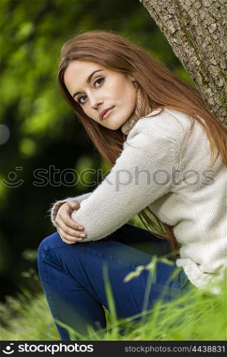 Outdoor portrait of beautiful thoughtful sad girl or young woman with red hair wearing a white jumper sitting &amp; leaning against a tree in the countryside