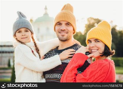 Outdoor portrait of friendly family stand close to each other, have broad smiles. Pretty woman in yellow trendy hat and red sweater, handsome young man holds little adorable daughter on hands